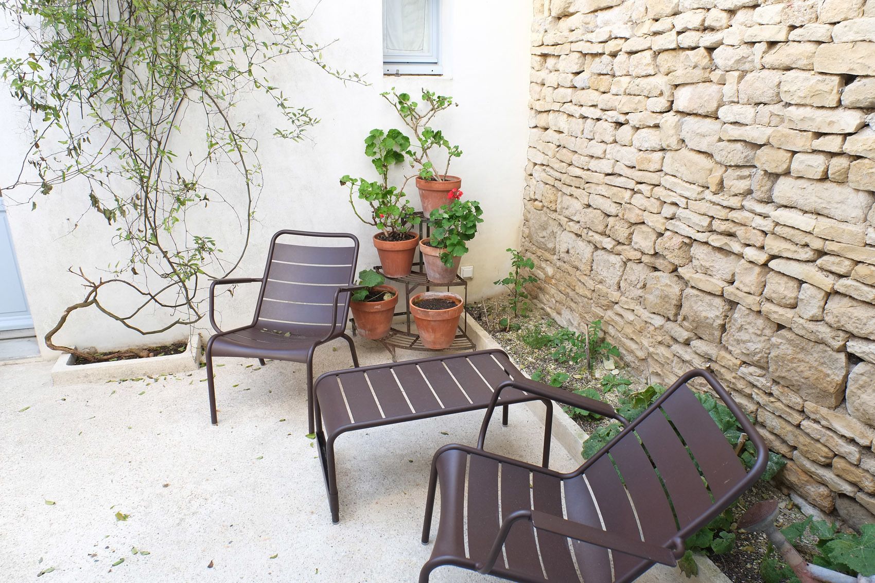 Zona d'estar a l'exterior amb dues cadires metàl·liques i una taula, envoltada de plantes en testos i una paret de pedra.