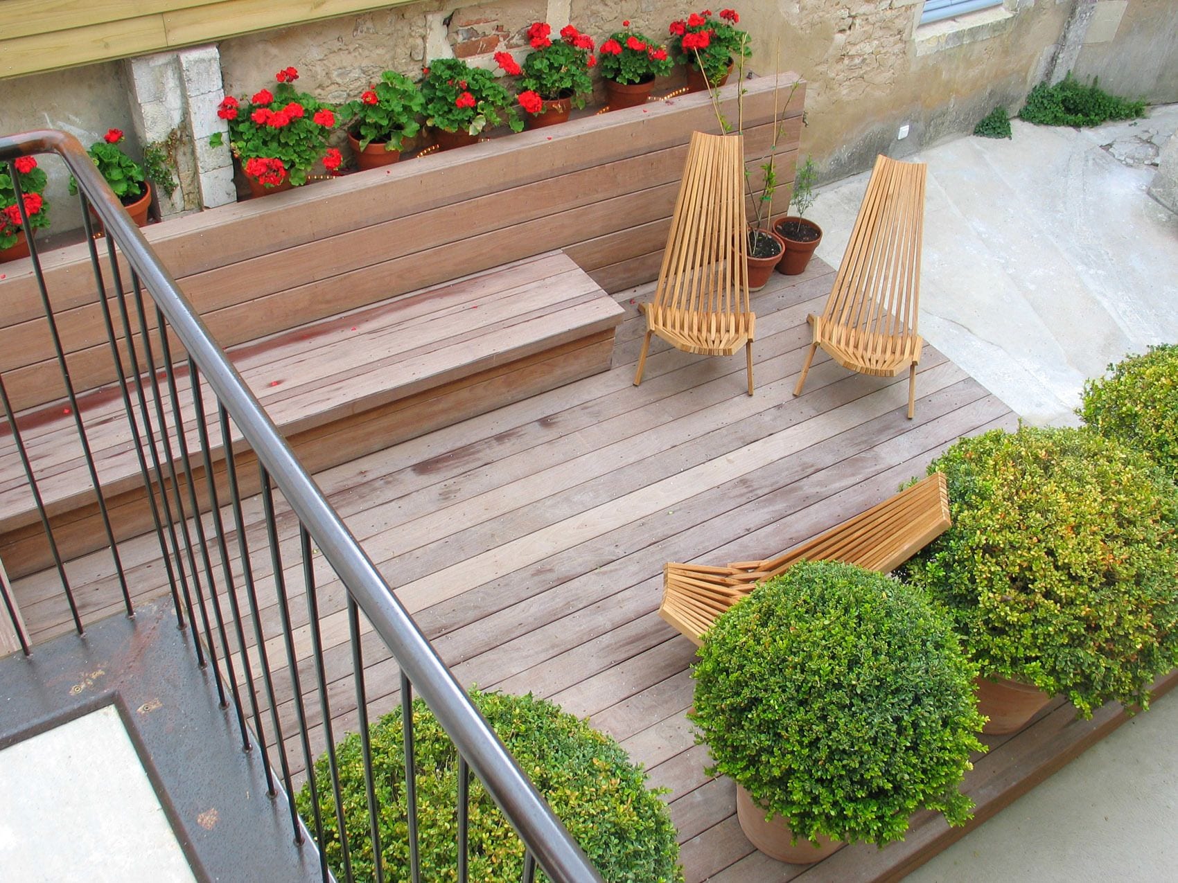 A wooden deck with steps leading up to a balcony, two wooden chairs, and potted plants.