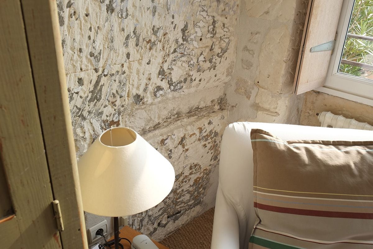 A room with a rustic, textured wall, a bedside lamp, a bed with a striped pillow, and a window with a view of greenery.