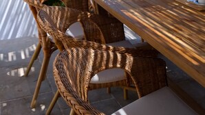Uma área de refeições ao ar livre com cadeiras de vime e uma mesa de madeira, com vista para uma piscina e vegetação.