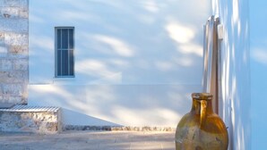 Uma trilha de pedra estreita conduz a um edifício branco com uma pequena janela e telhado de pedra. Um vaso rústico grande está colocado no chão à direita.