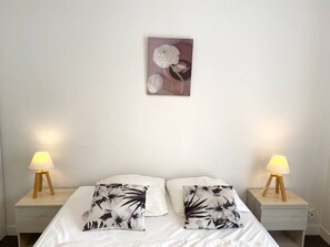 A neatly made bed with white bedding and two floral-patterned pillows. There are two bedside tables with lamps, and a framed floral artwork is hung on the wall above the bed. - Spacious apartment with WIFI, 2 bedrooms, downtown Perros-Guirec, near beaches (Perros-Guirec)