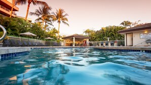 Une piscine entourée de palmiers et d’un bâtiment doté d’une zone abritée, avec des chaises longues et des parasols à proximité.