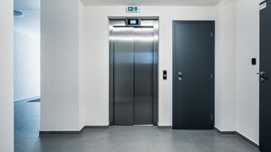An elevator in a hallway with a digital display showing the floor number, a door with a handle, and a wall-mounted emergency exit sign. - Smart & Green Living by Ambiente (Bratislava)