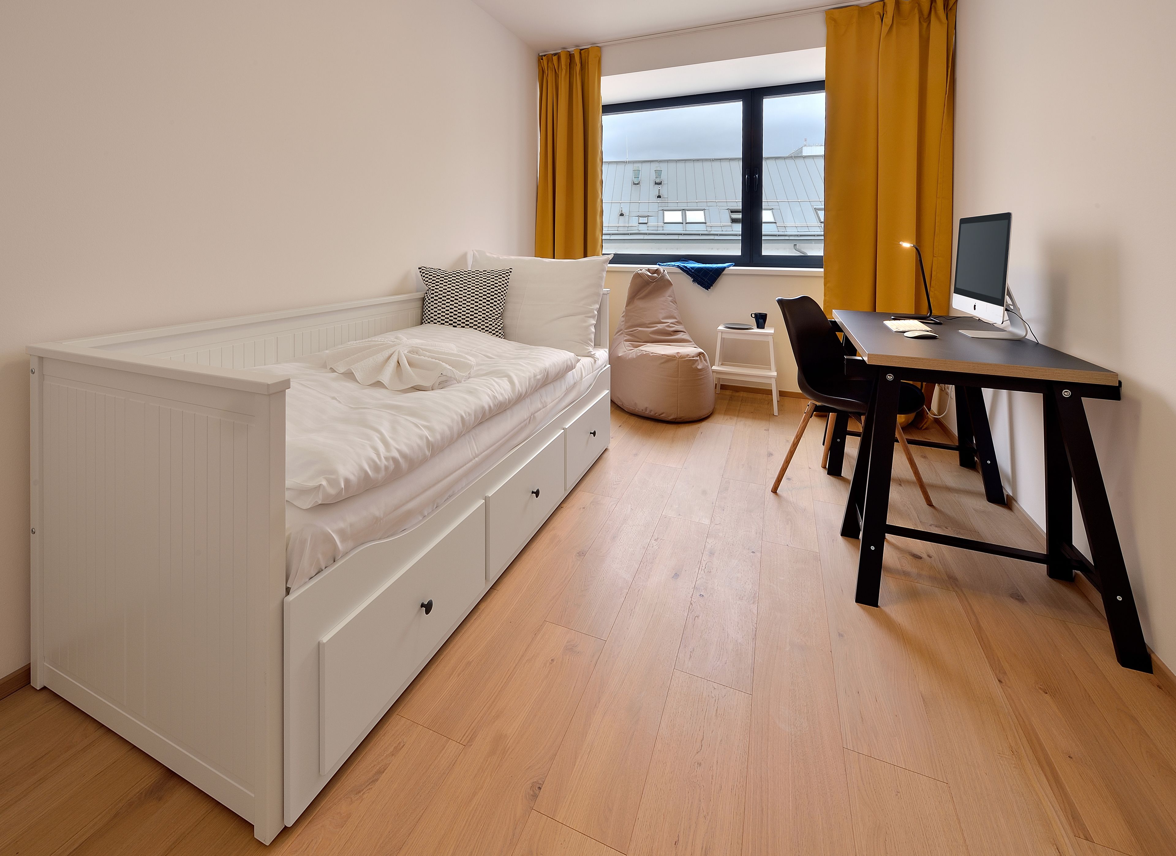 Un dormitorio con cama, escritorio, silla, sillón de mimbre y ventana con cortinas.