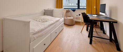 Un dormitorio con cama, escritorio, silla, sillón de mimbre y ventana con cortinas.