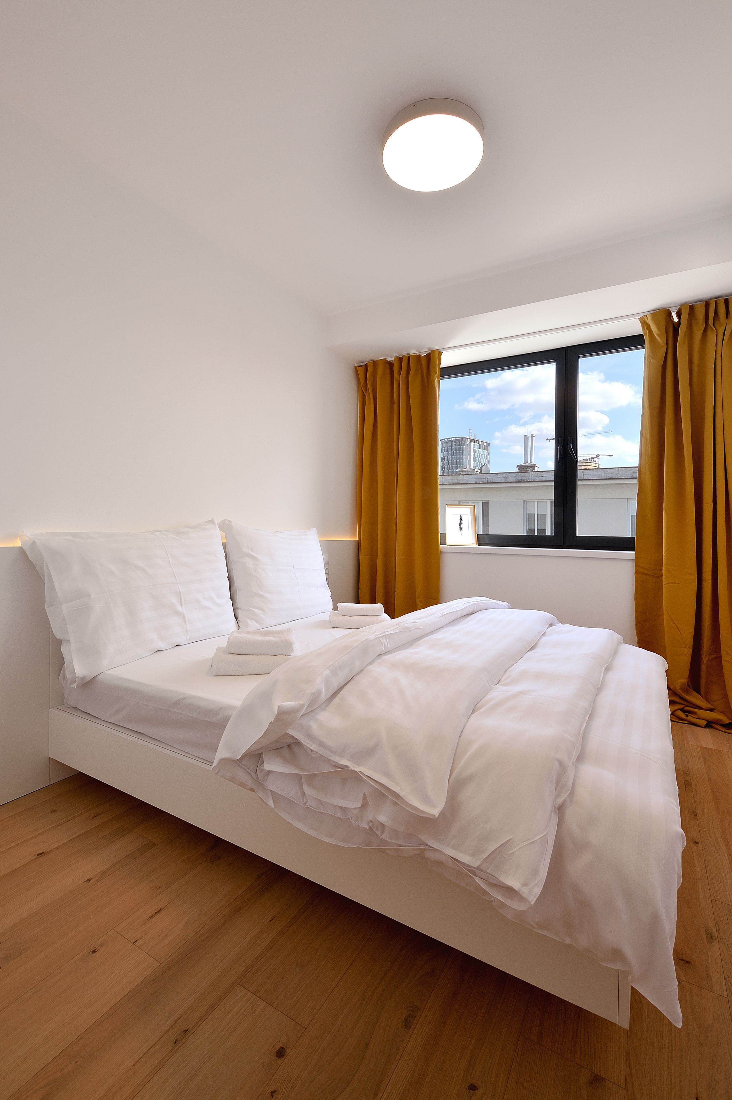 A neatly made bed with white linens in a room with wooden flooring, a window with yellow curtains, and a view of buildings outside.