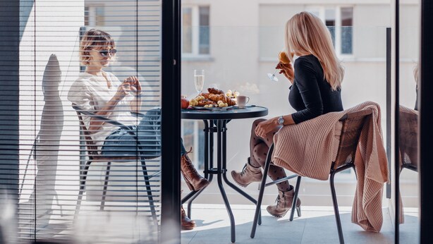 A person sitting at a table with a plate of food and a glass of water, in a room with large windows and a view of buildings outside. - Smart & Green Living by Ambiente (Bratislava)