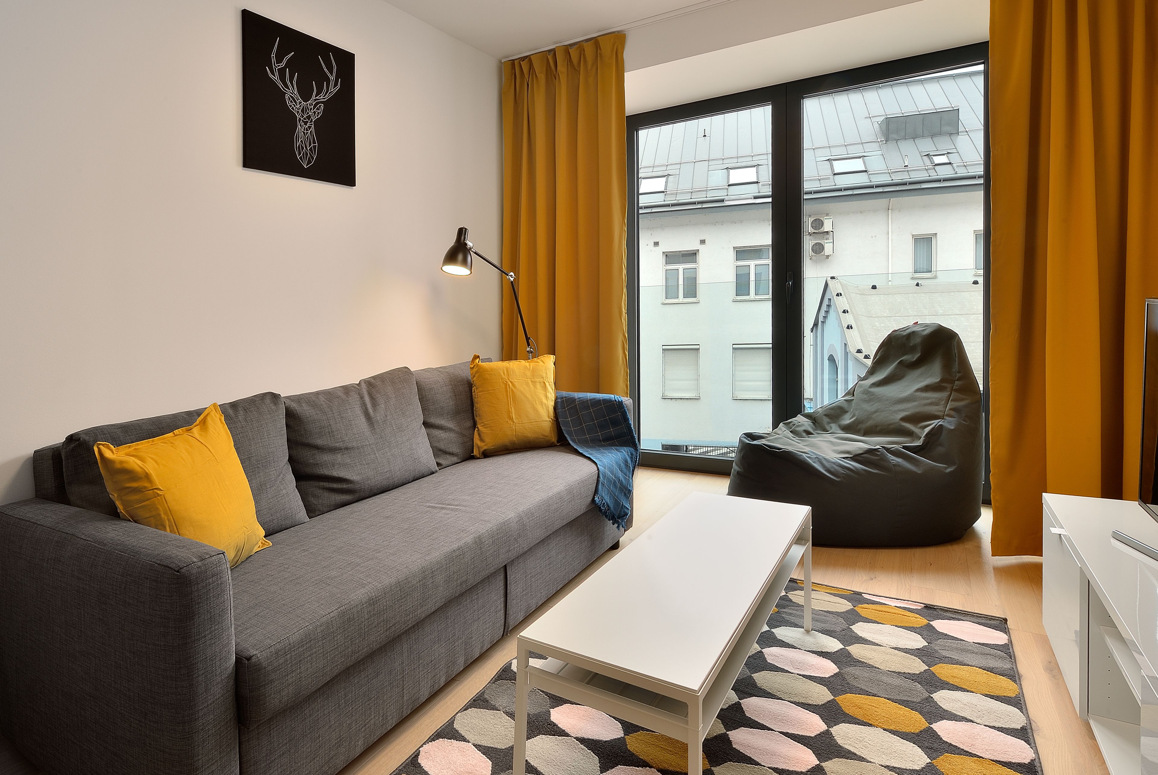 Een moderne woonkamer met een grijze bank, gele kussens, een witte salontafel, een relaxfauteuil en een groot raam met gele gordijnen.