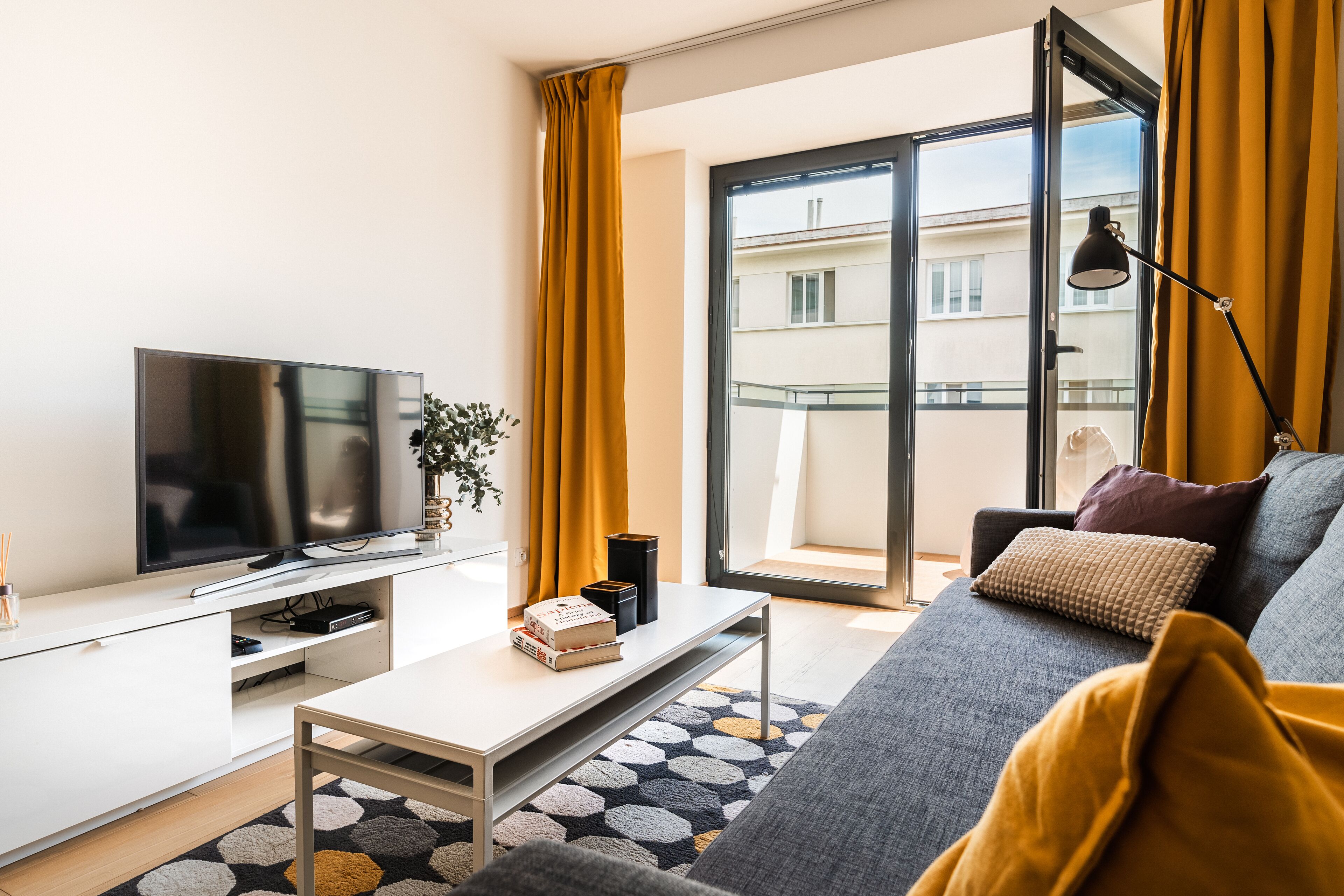 A modern living room with a flat-screen TV, a coffee table, a sofa, and a large window with yellow curtains.