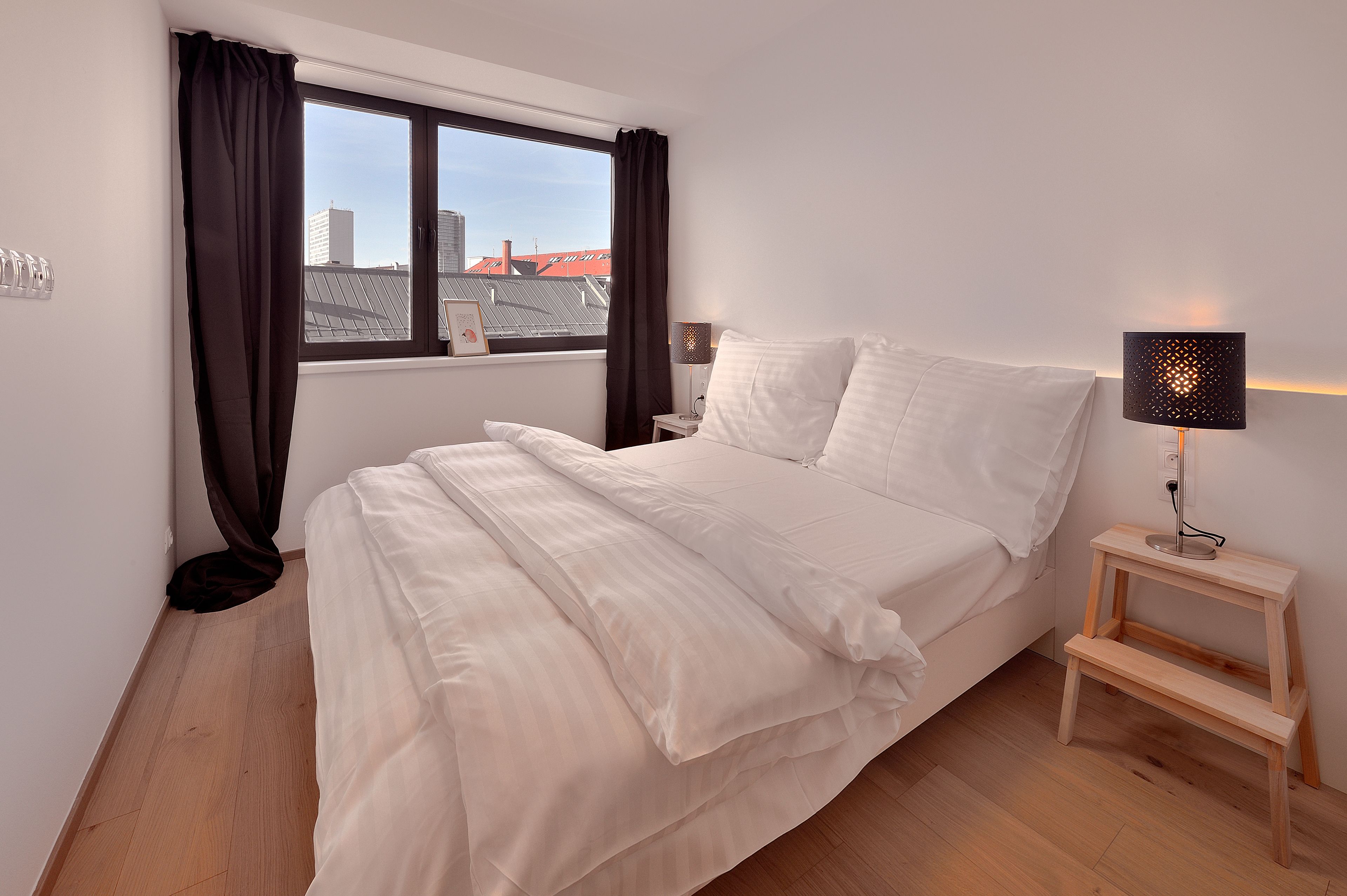 A neatly made bed with white linens, a bedside lamp, and a view of buildings through a window with dark curtains.