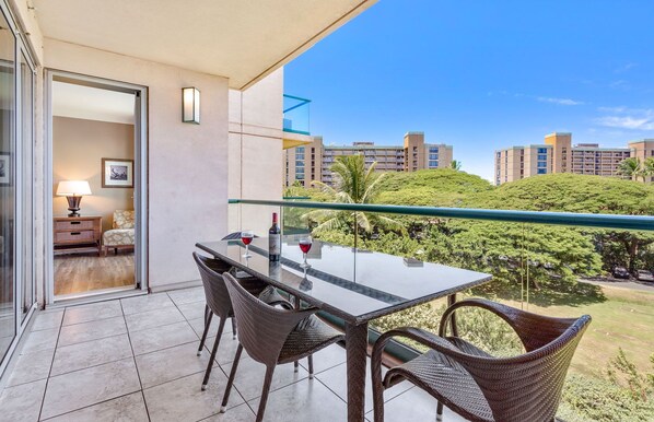 A balcony with a glass table and wicker chairs, overlooking a lush green landscape and a cityscape.