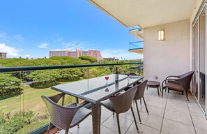 A balcony with a glass table and wicker chairs, overlooking a green landscape and buildings.