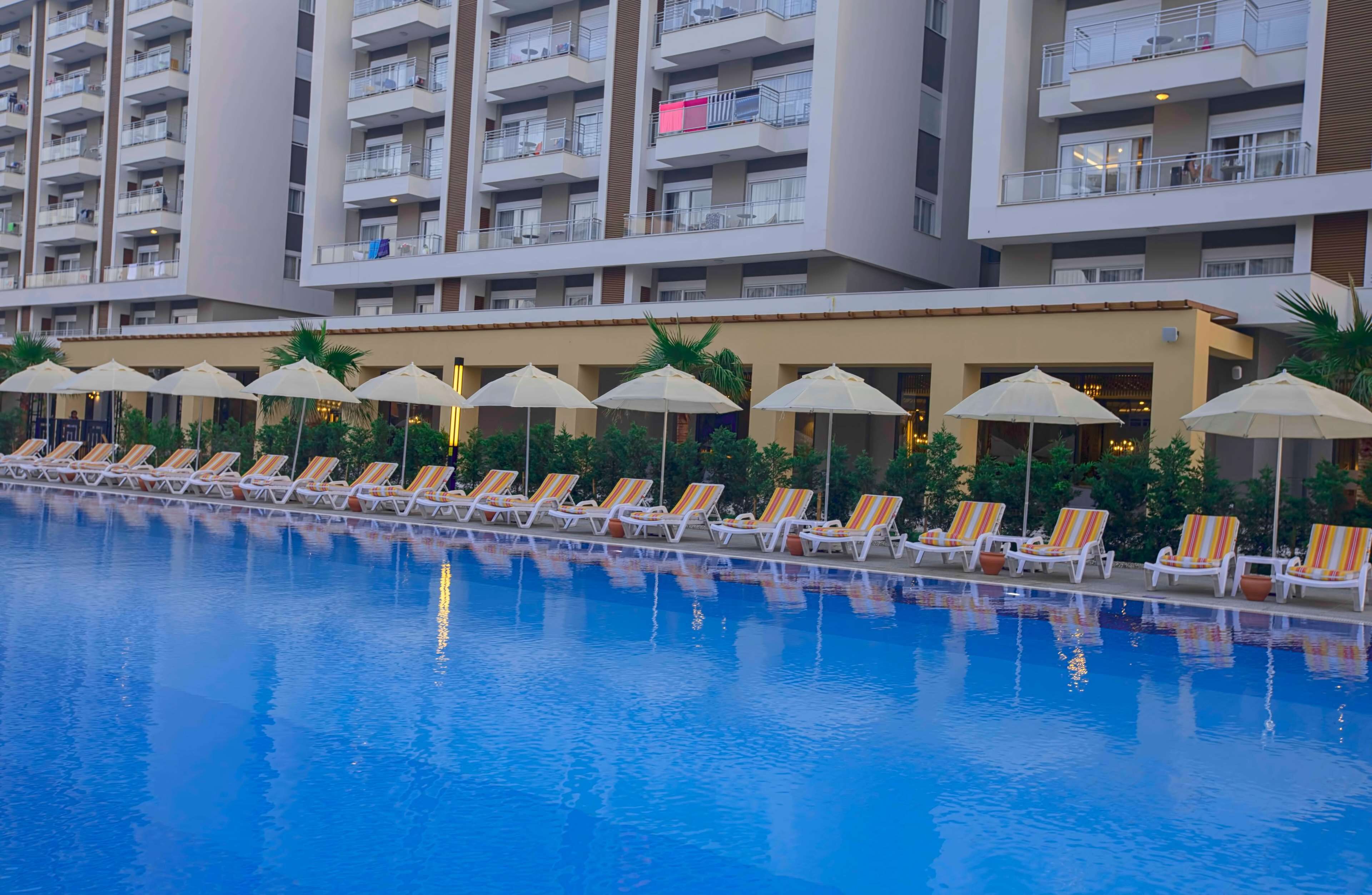 Una piscina con lettini e ombrelloni, circondata da un edificio a più piani con balconi.
