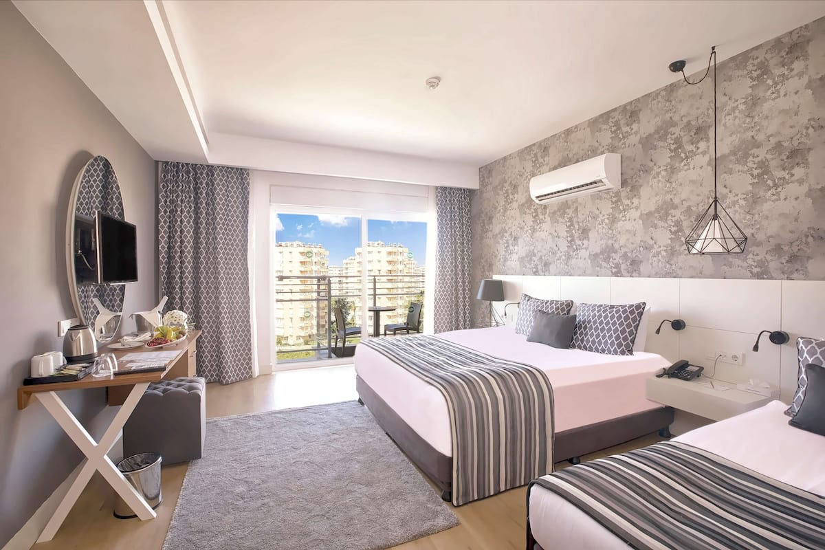 Une chambre d’hôtel moderne avec deux lits, un bureau avec un miroir, une petite table et un balcon offrant une vue sur les bâtiments.