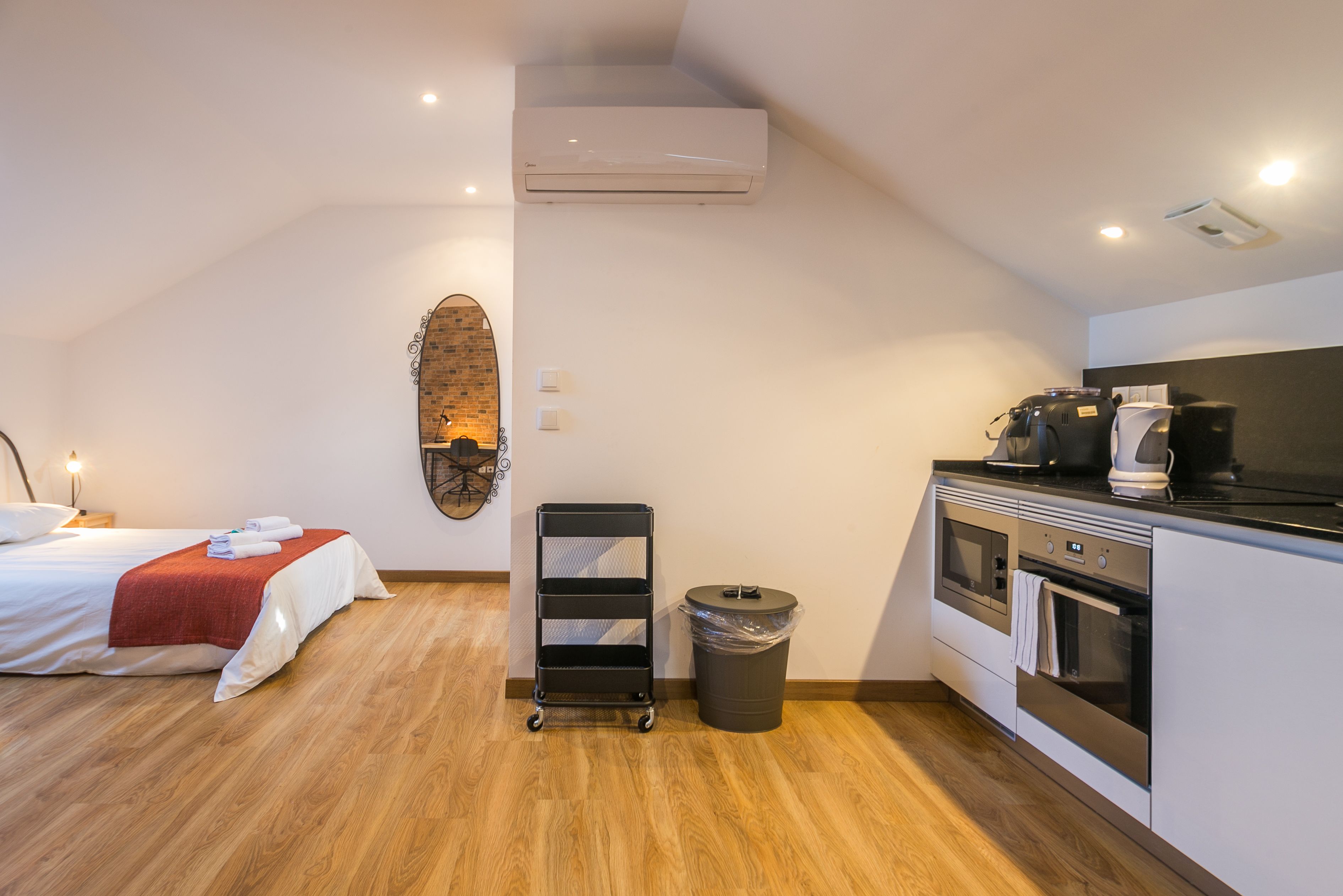 A hotel room with a bed, a small kitchenette, a trash bin, and a wall-mounted shelf.