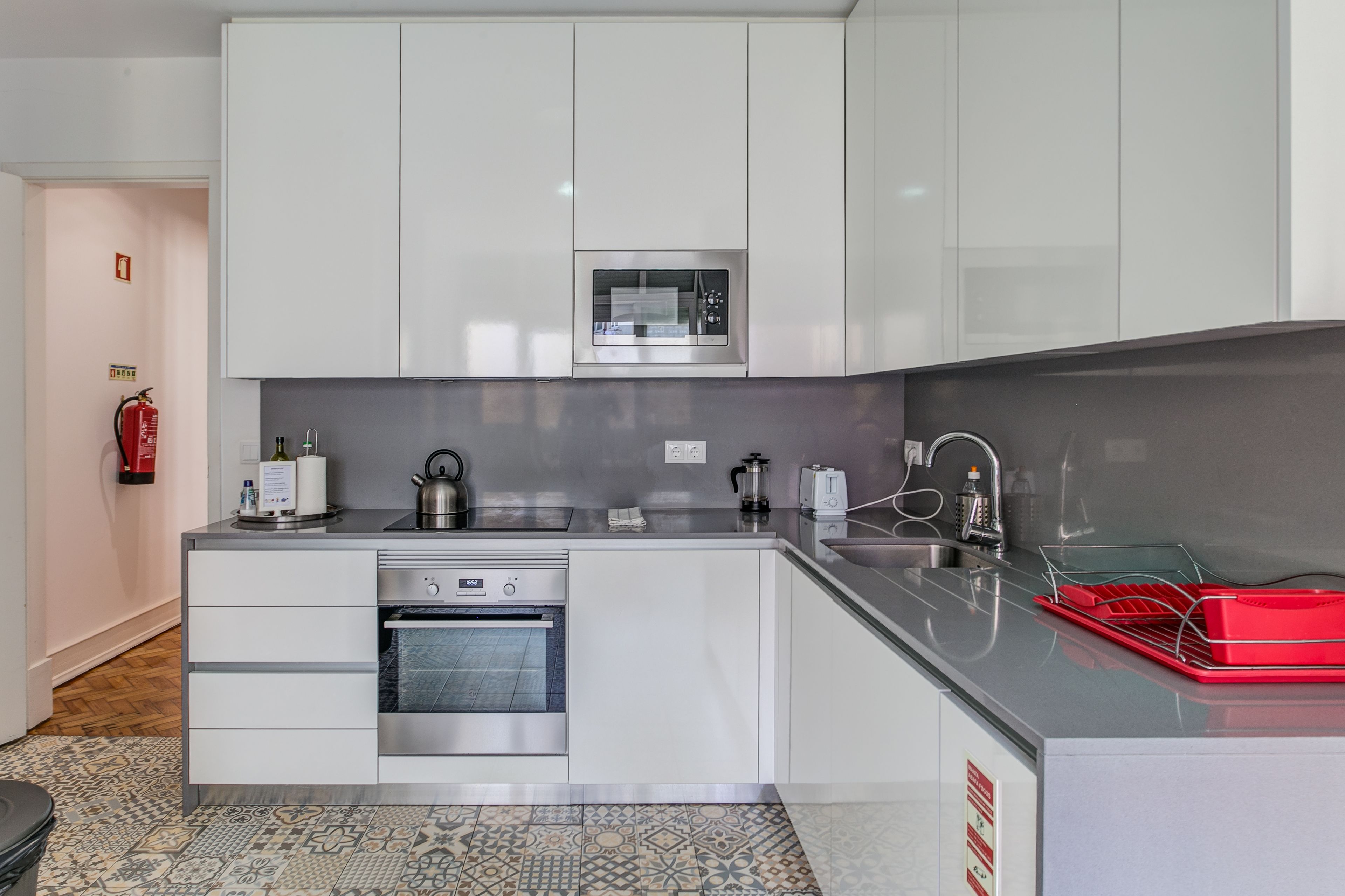 A modern kitchen with white cabinets, a built-in microwave, a kettle, and a red dish rack.