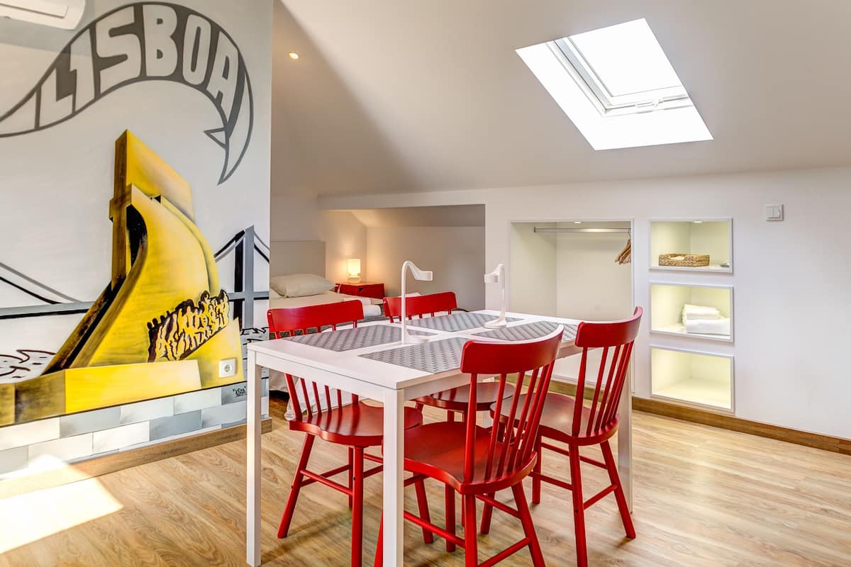 Une pièce moderne avec un coin repas comprenant une table blanche et des chaises rouges, une grande œuvre d’art au mur et une lucarne.