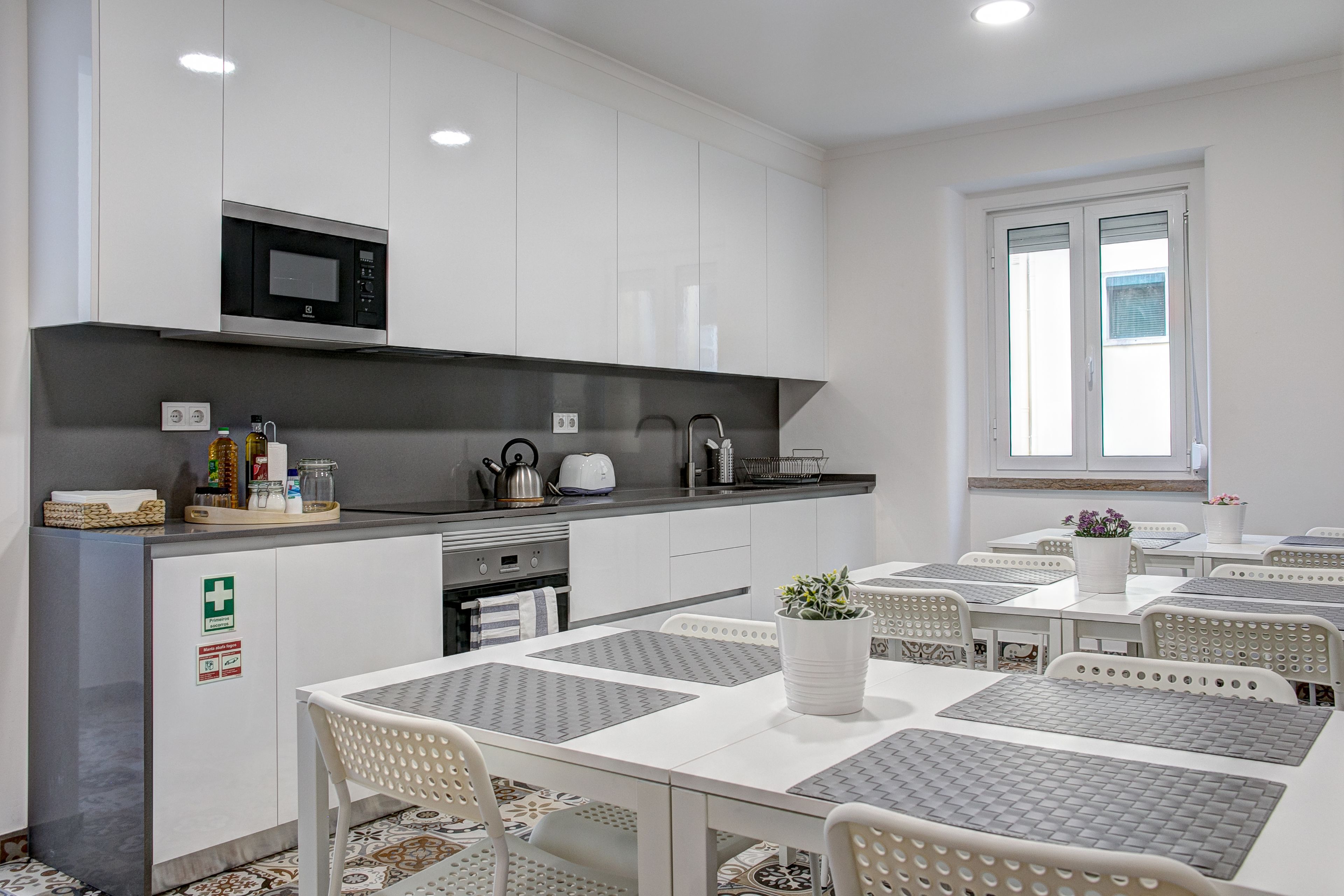 Una cocina moderna con armarios blancos, un microondas y una tetera en la encimera. Hay dos ventanas con persianas, y un comedor con sillas y una mesa blancos.