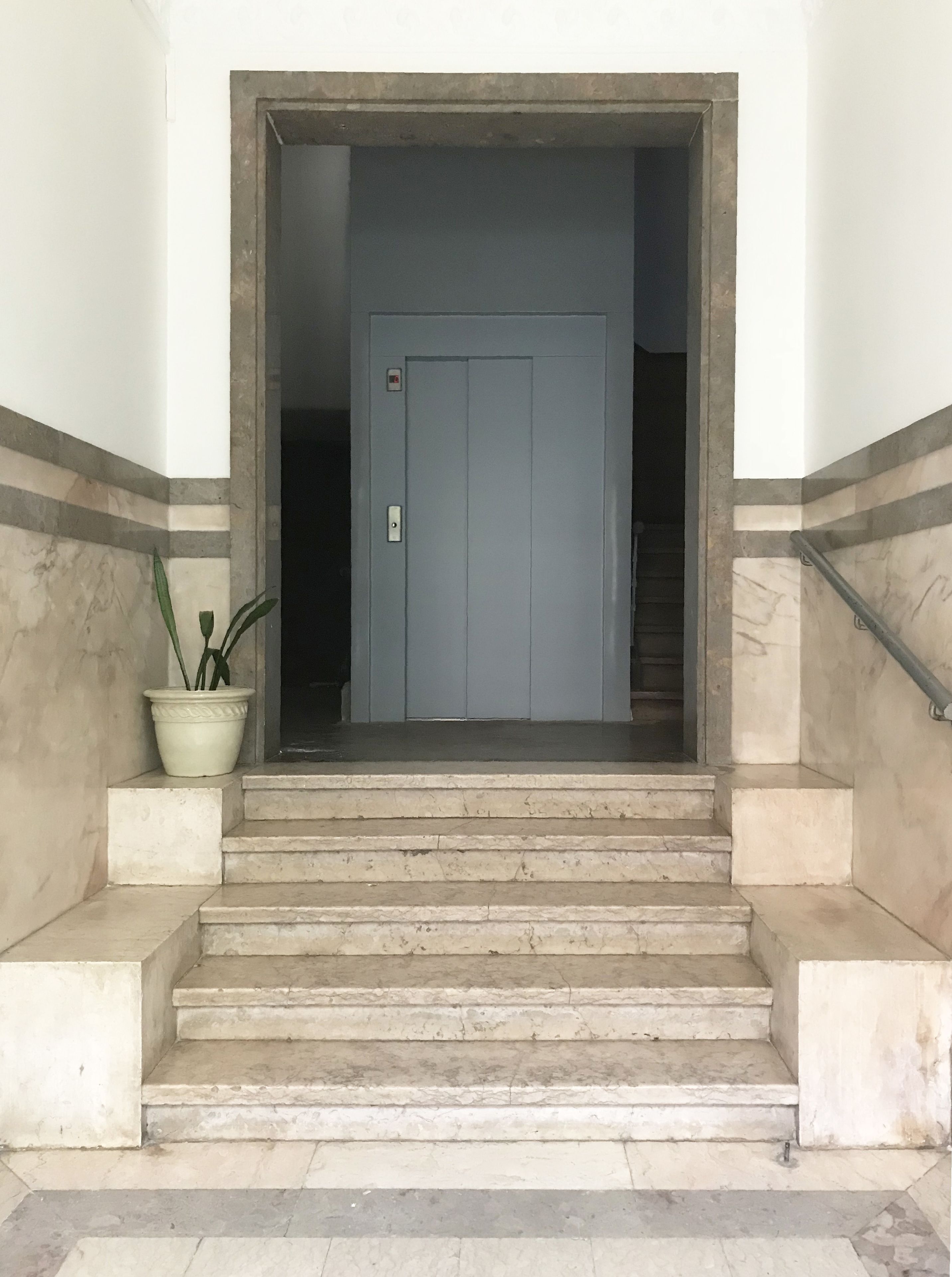 Una serie di gradini in marmo che conducono a una porta dal design semplice, incorniciata da pareti decorate con un motivo marmorizzato.