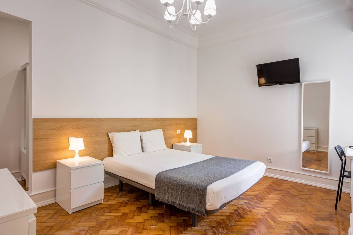 Une chambre à coucher avec une tête de lit en bois, un lit recouvert de draps blancs, deux tables de chevet avec des lampes, un téléviseur à écran plat fixé au mur et un miroir.