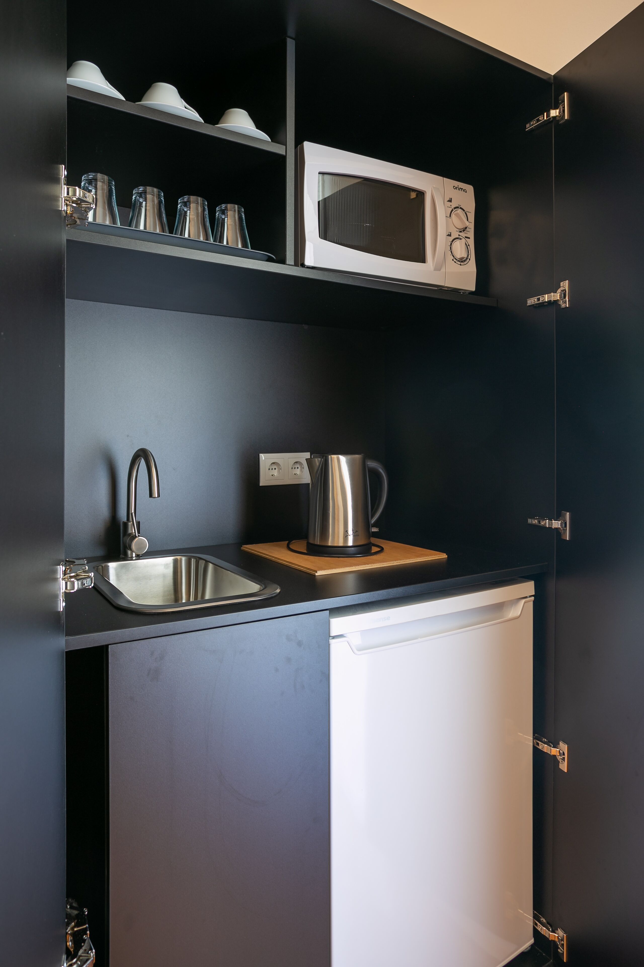 Una cocina compacta con microondas integrado, un lavamanos de acero inoxidable y un hervidor sobre la encimera.