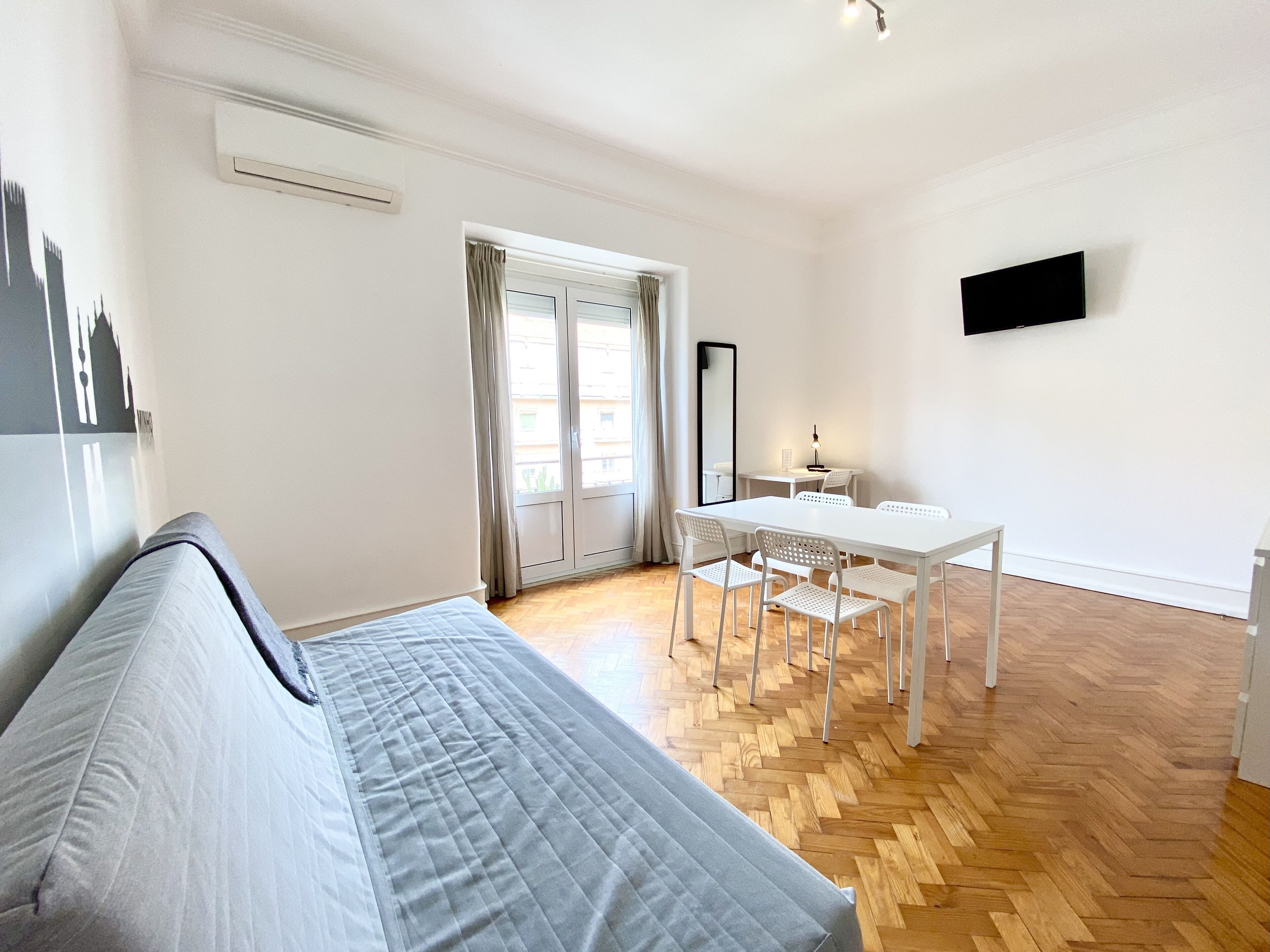 Une chambre moderne et bien éclairée, comprenant un lit, une table à manger avec des chaises, un téléviseur à écran plat fixé au mur et une grande fenêtre avec des rideaux.
