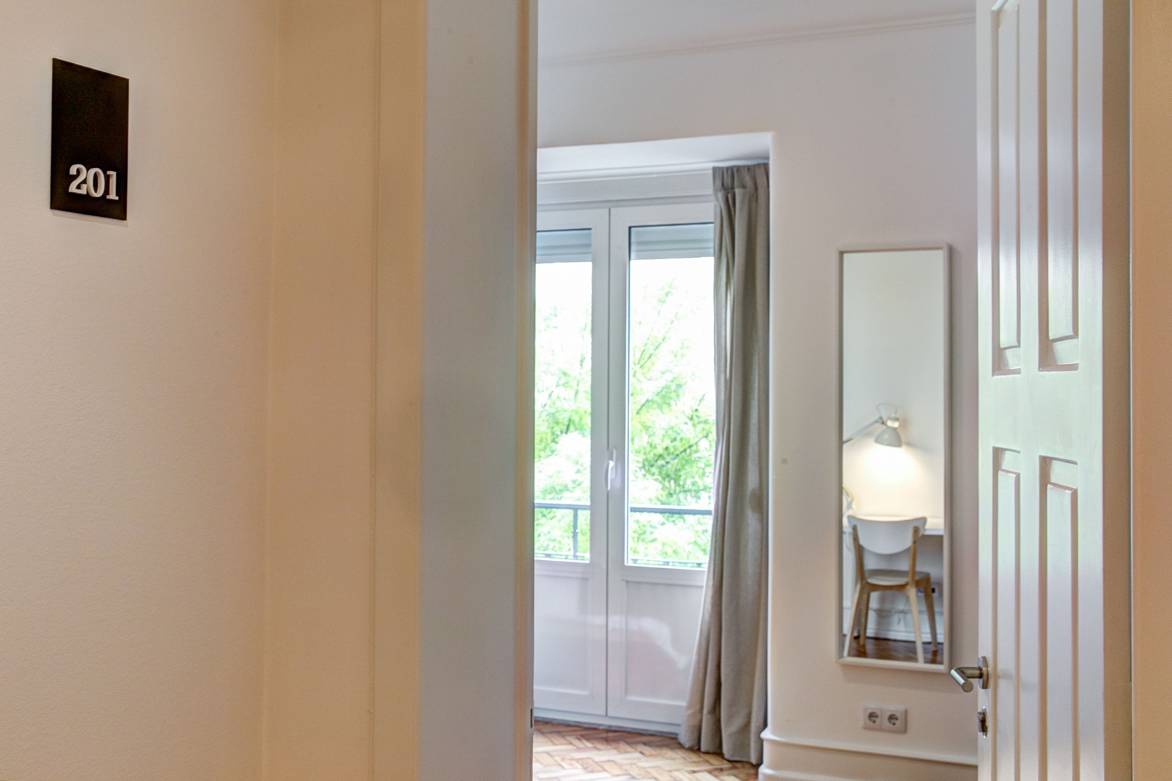 Habitación de hotel con una puerta identificada con el número 201, una ventana con cortinas y una lámpara sobre una mesa.