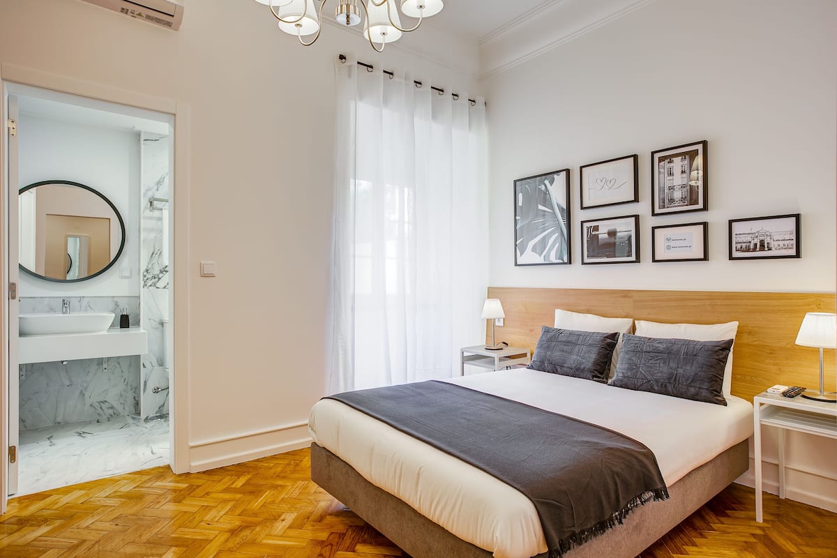 Une chambre avec un grand lit, une table de chevet avec une lampe, une commode, un miroir et une fenêtre avec des rideaux légers.