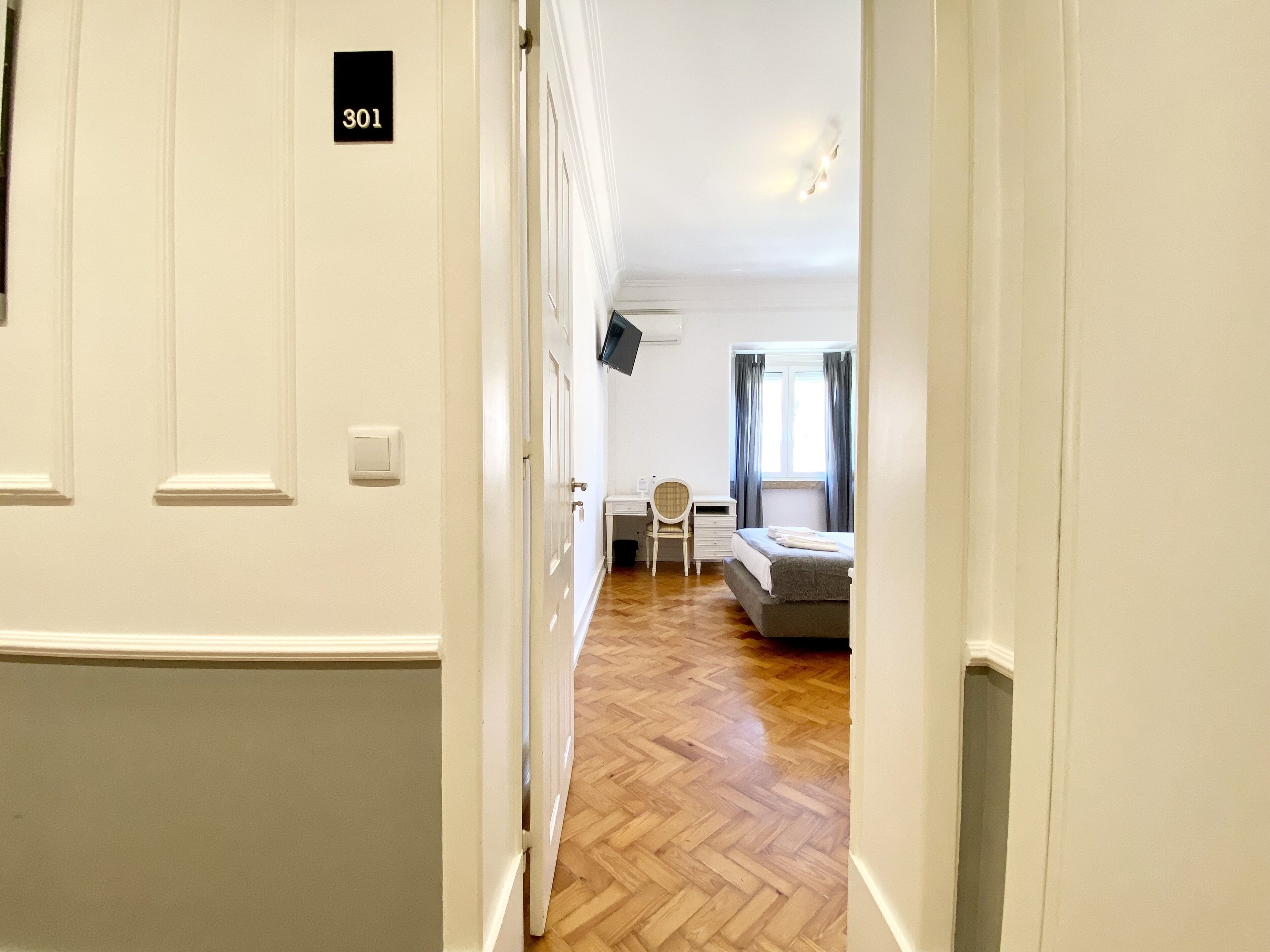 Corredor que dá para um quarto com cama, escrivaninha e cadeira.
