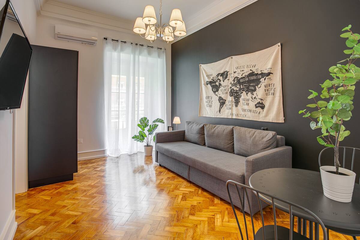Un salon moderne avec un canapé gris, un parquet en bois, une tapisserie représentant une carte du monde, une plante en pot et une table à manger avec des chaises.