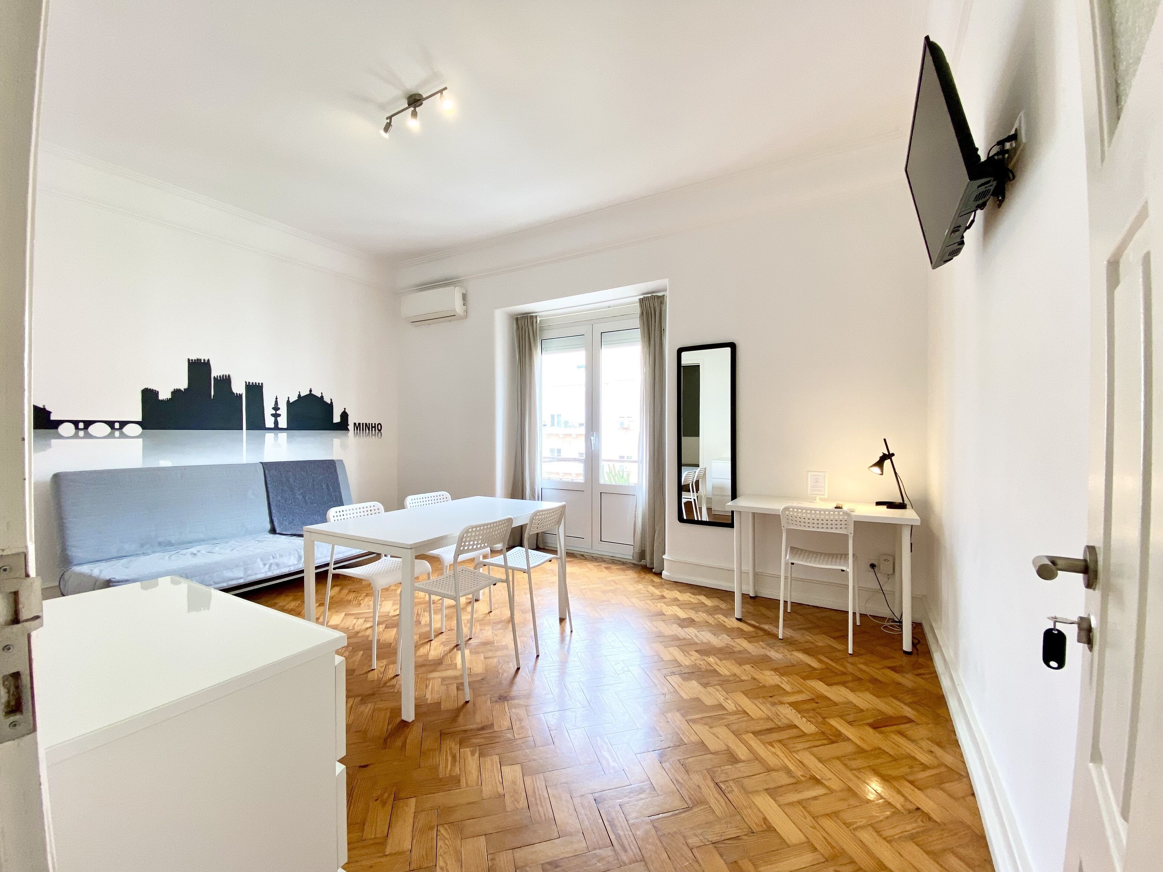A modern, well-lit room with a wooden herringbone floor, white furniture, a flat-screen TV mounted on the wall, and a cityscape mural.