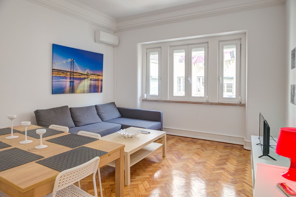 Un salon moderne avec un canapé, une table à manger et des chaises. Un tableau représentant un pont est accroché au mur, il y a une fenêtre avec des rideaux et une lampe rouge posée sur une table.