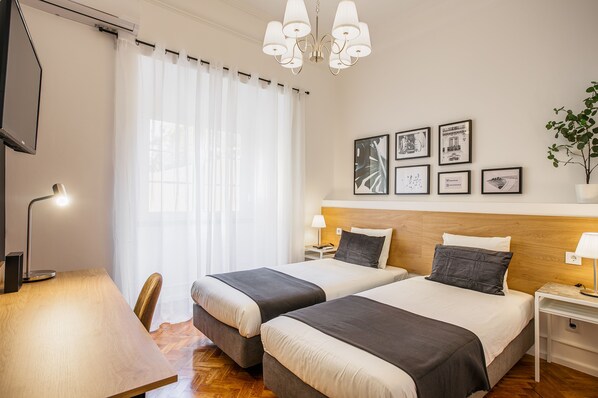 Quarto de hotel com duas camas, uma cabeceira de madeira, uma escrivaninha, uma cadeira, uma televisão, um candelabro e quadros nas paredes.