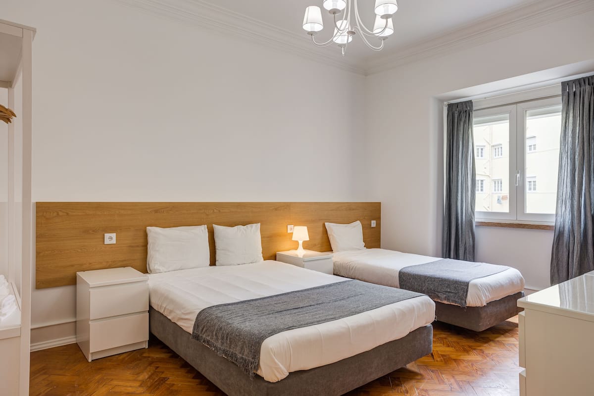 Une chambre d’hôtel avec deux lits, une tête de lit en bois, une table de chevet avec une lampe et une fenêtre avec des rideaux.