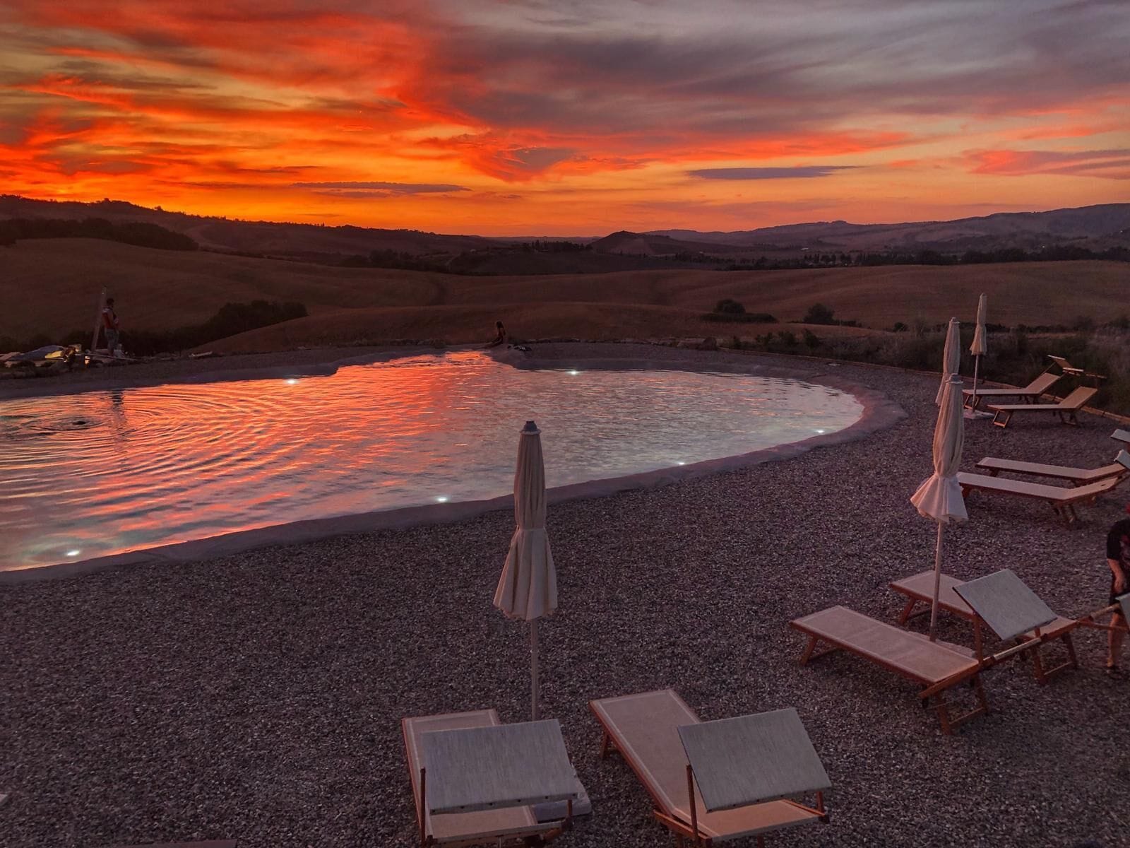 Ein Kiesstrand mit Liegestühlen und Sonnenschirmen, ein Pool, der die Farben des Sonnenuntergangs reflektiert, und sanfte Hügel im Hintergrund.
