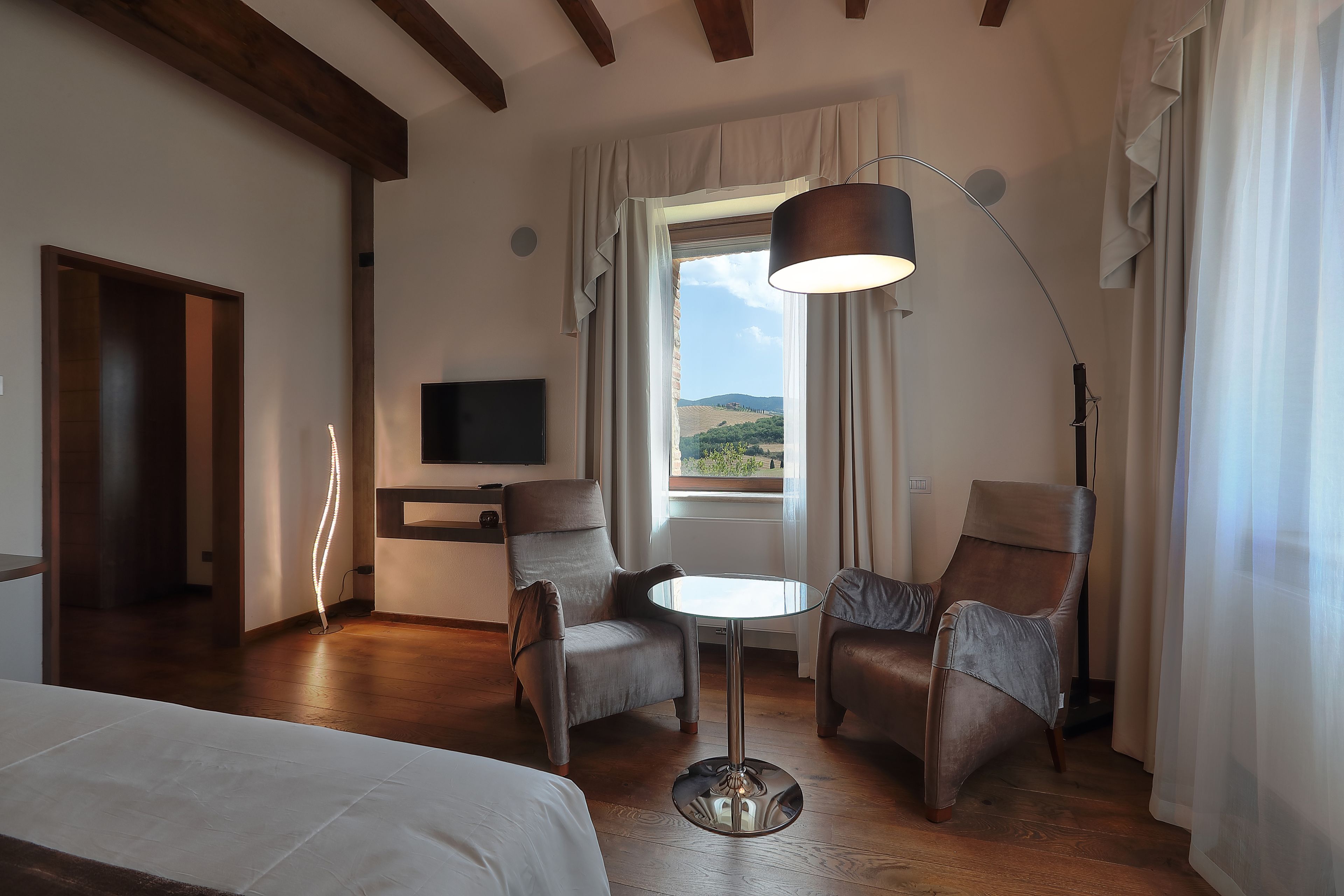 A hotel room with a bed, two armchairs, a small round table, a TV, and a window with a view of the countryside.