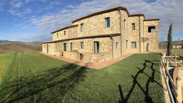 A stone building with multiple windows and a flat roof, surrounded by a well-maintained lawn and a hilly landscape in the background.
