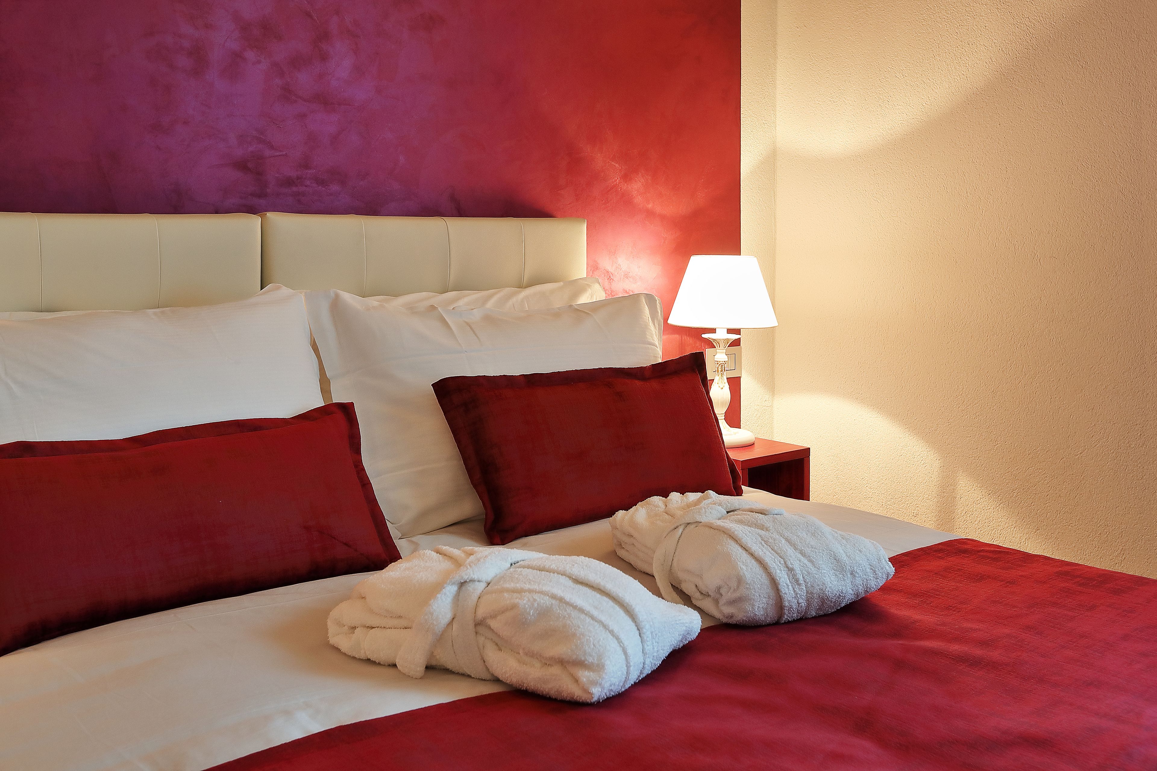 Quarto de hotel com uma cama, almofadas vermelhas e brancas, um cobertor vermelho e dois roupões de banho brancos.
