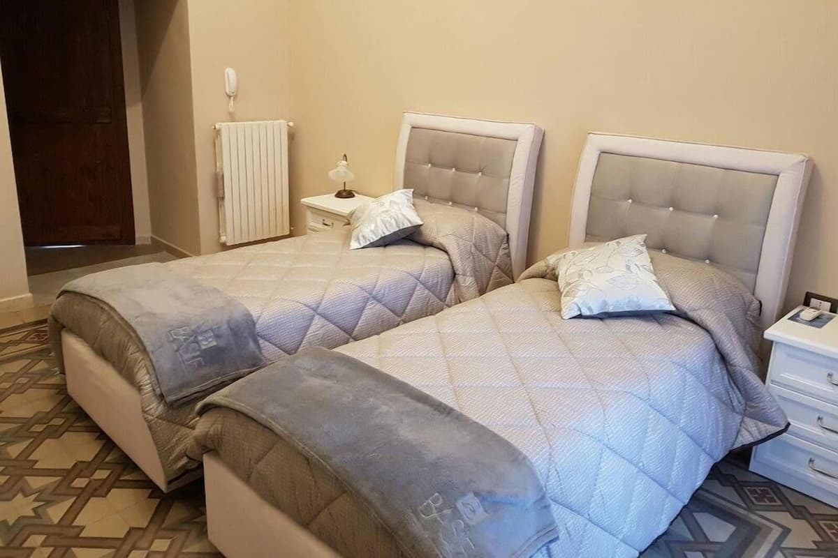 Un dormitorio con dos camas, una mesita de noche con lámpara, un radiador y un suelo estampado.