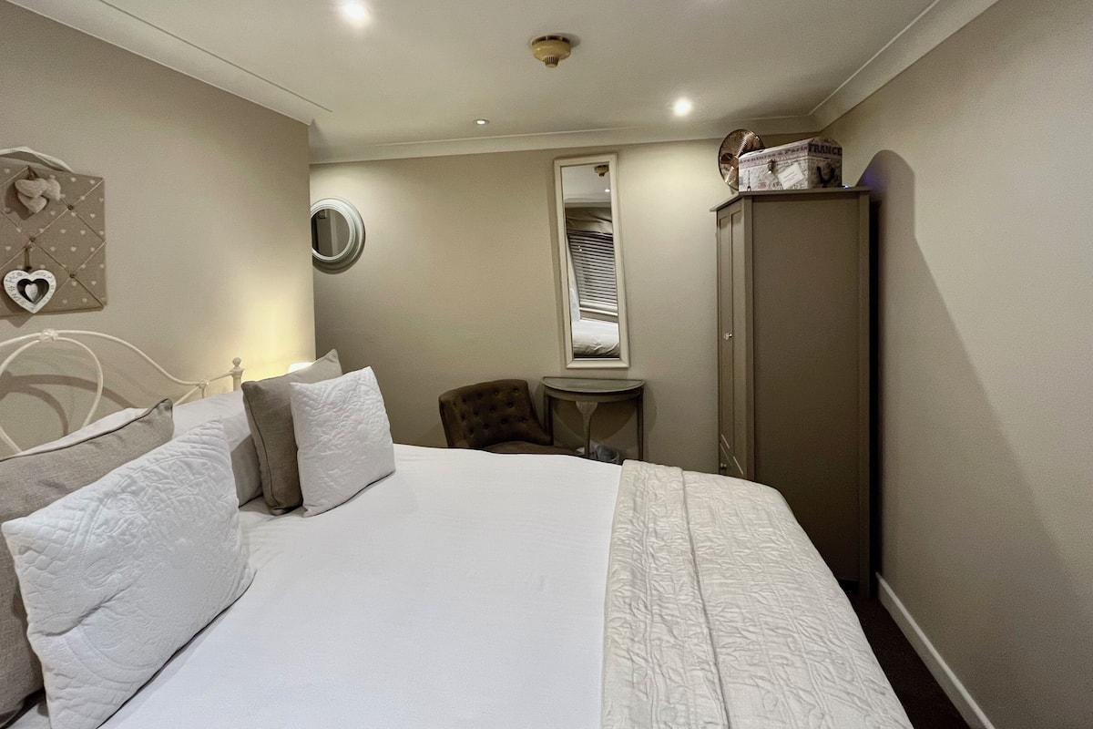 A hotel room with a bed, a chair, a small table, a wardrobe, and a mirror.