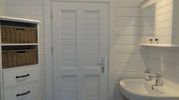 Un baño con una puerta blanca, cestas de mimbre en un mueble, un lavamanos y un espejo encima.
