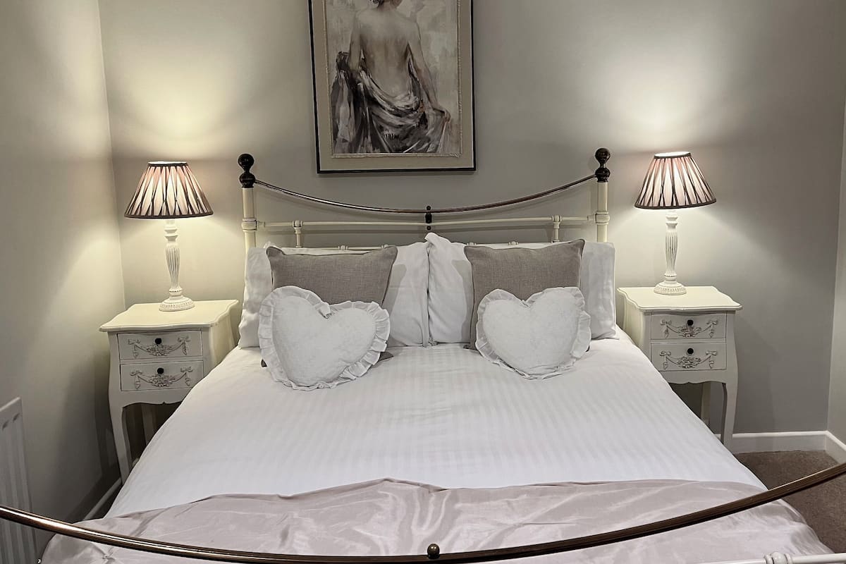 A neatly made bed with white linens and heart-shaped pillows, flanked by two bedside tables with lamps, in a room with a minimalist design and a painting above the bed.