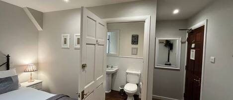 Un dormitorio con cama, mesita de noche con lámpara, y puerta que da a un baño con inodoro, lavabo y espejo.