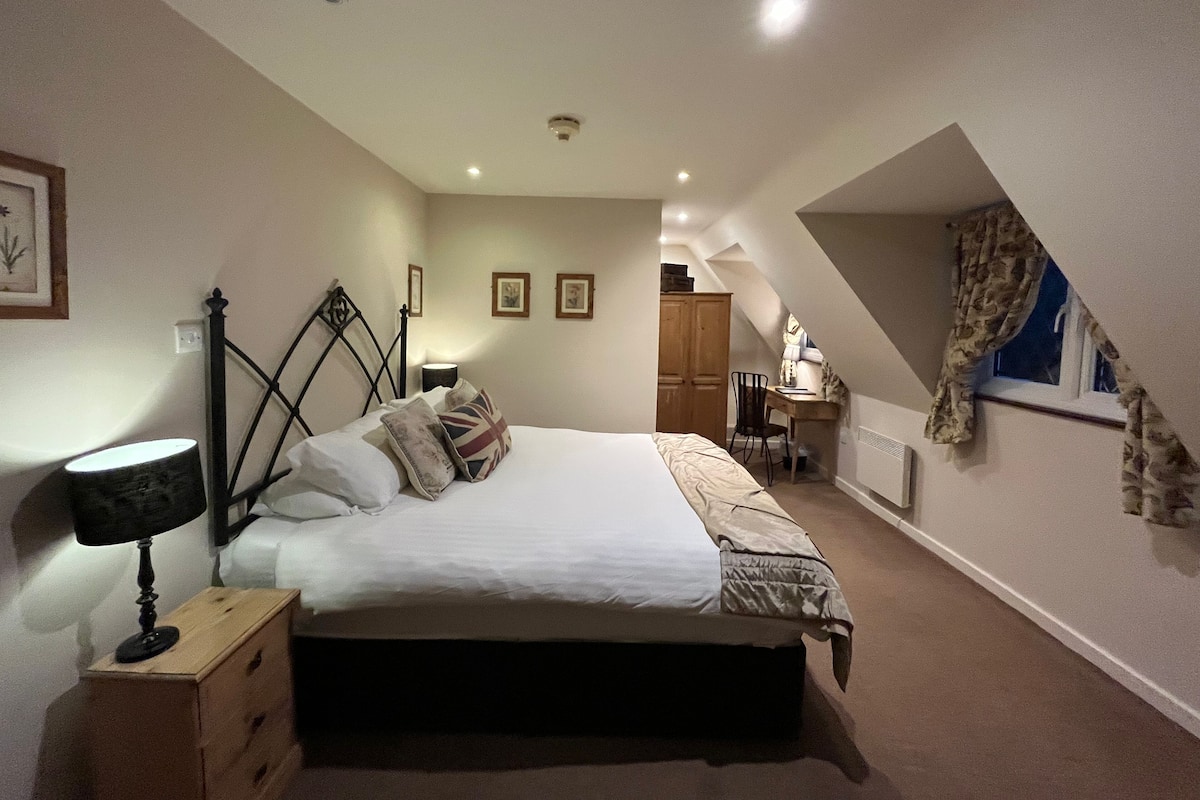 A bedroom with a bed, bedside table, lamp, and a desk with a chair. There is a window with curtains and a radiator on the wall.