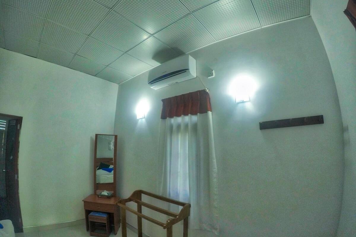 A small, well-lit room with a bed, a bedside table, a mirror, and a window with curtains.