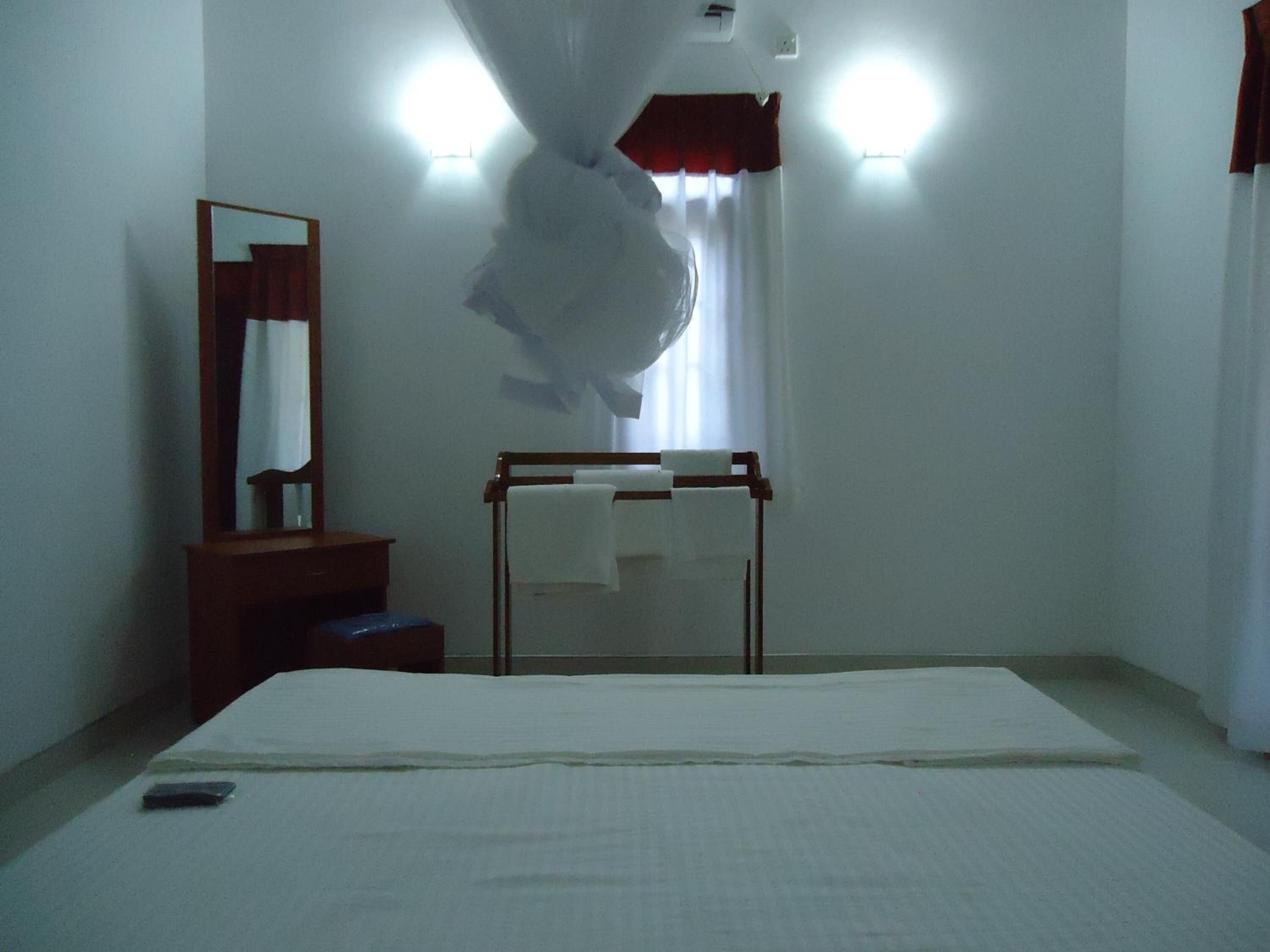 A hotel room with a bed, a bedside table, a mirror, a hanging shower curtain, and a window with curtains.