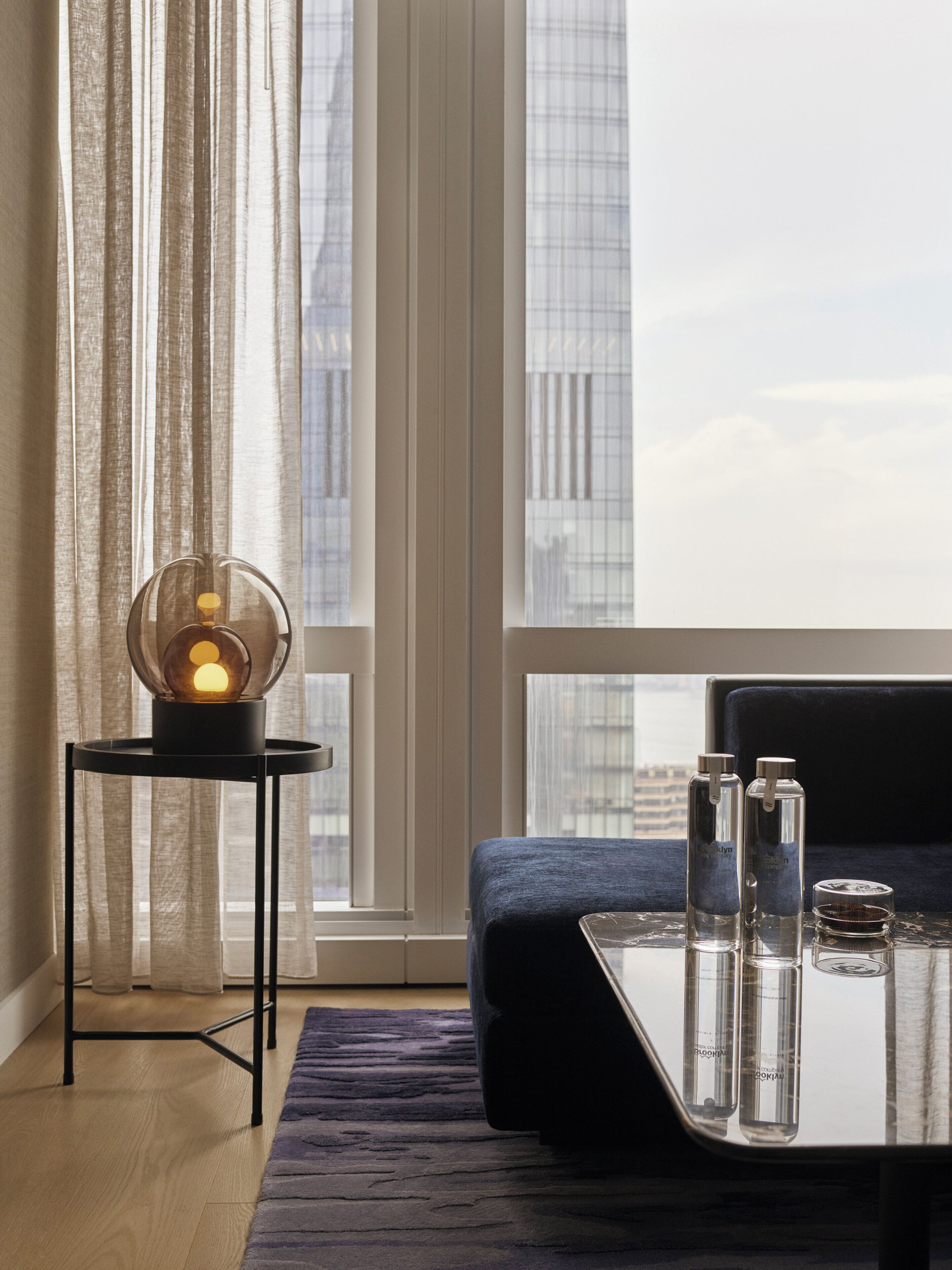 Una habitación moderna con vistas despejadas de la ciudad a través de amplios ventanales, una mesa redonda con una lámpara esférica, un sofá azul y una mesa de centro de cristal con botellas.