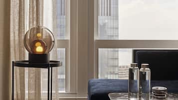 A modern room with a clear view of the cityscape through large windows, a round table with a spherical light, a blue sofa, and a glass coffee table with bottles.