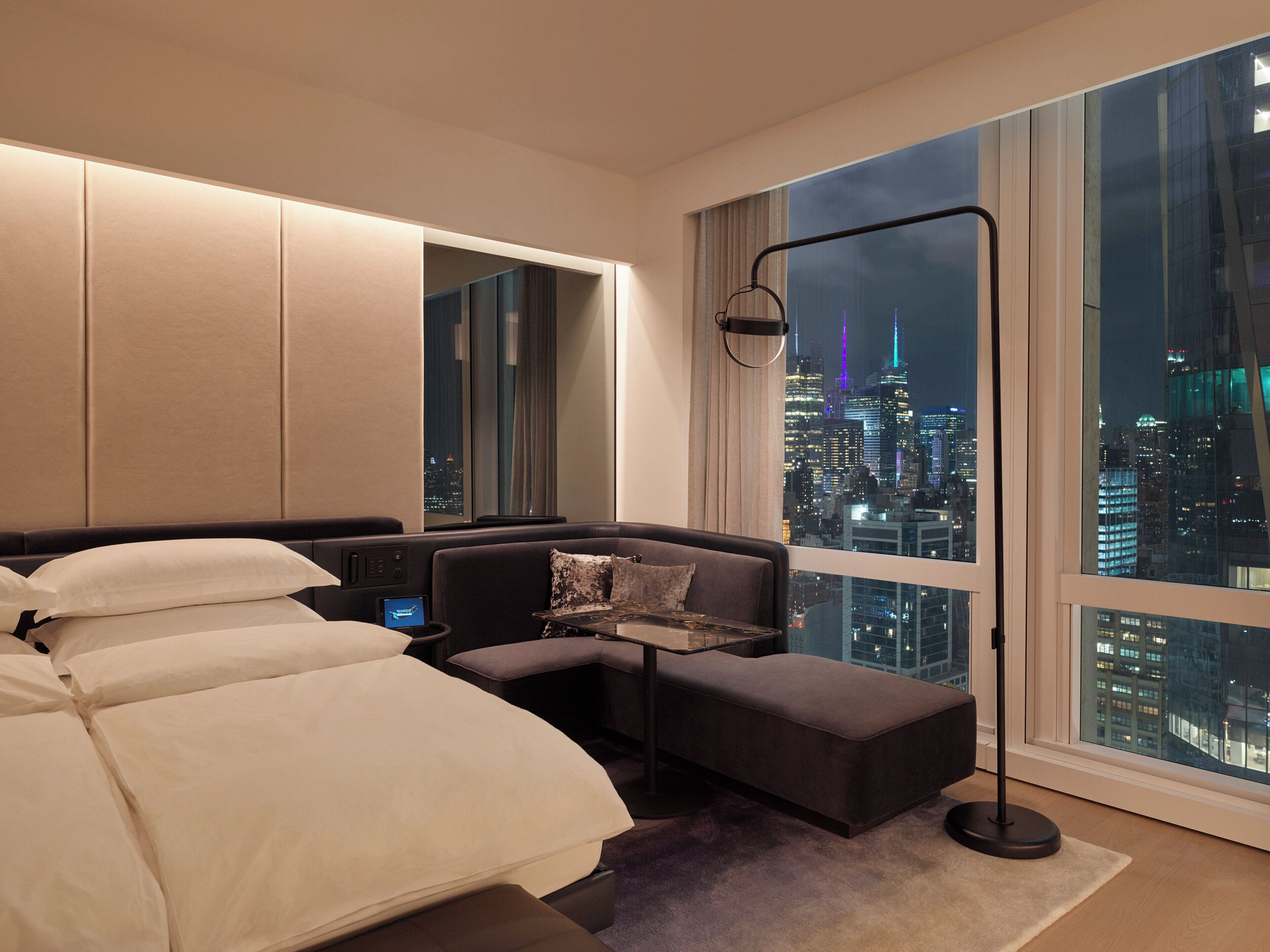 A modern hotel room with a large bed, a seating area, a desk, and a view of a cityscape at night.